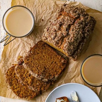 An overhead view of a loaf of best homemade pumpkin bread that has three slices cut from it and resting on brown paper with two glass mugs of creamed coffee nearby.