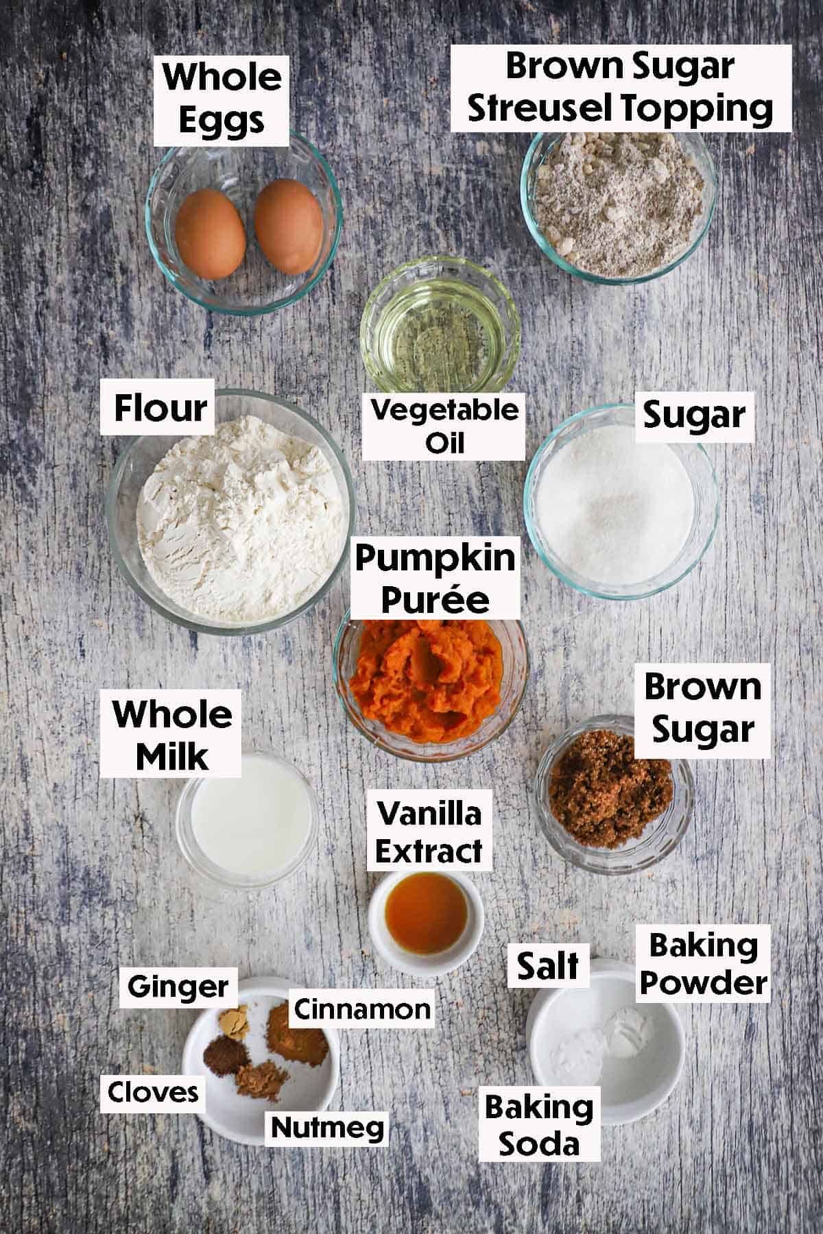 An arrangement of ingredients for best homemade pumpkin bread on a grey wooden background including pumpkin purée, eggs, flour oil, sugar, milk, vanilla, and spices.