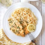 An overhead view of an elegant white dinner plate filled with a serving of homemade chicken piccata with a pile of linguine and a lemon wedge off to the side.