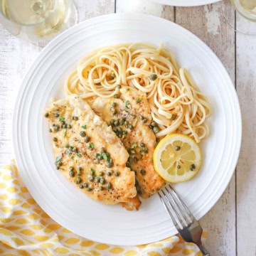 An overhead view of an elegant white dinner plate filled with a serving of homemade chicken piccata with a pile of linguine and a lemon wedge off to the side.