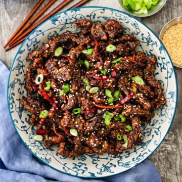 An overhead view of a colorful serving bowl filled with crispy chili beef garnished with slivers of red peppers, chopped scallions, and toasted sesame seeds.