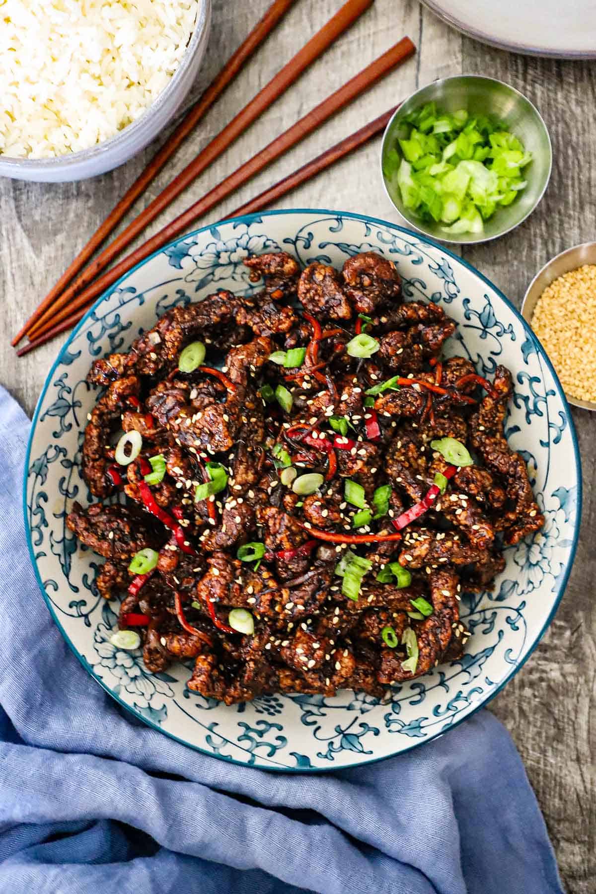 An overhead view of a colorful serving bowl filled with crispy chili beef garnished with slivers of red peppers, chopped scallions, and toasted sesame seeds.
