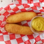 Two homemade corn dogs next to each other with a small container of yellow mustard nearby all on a red and white checkered wax paper in a basket.