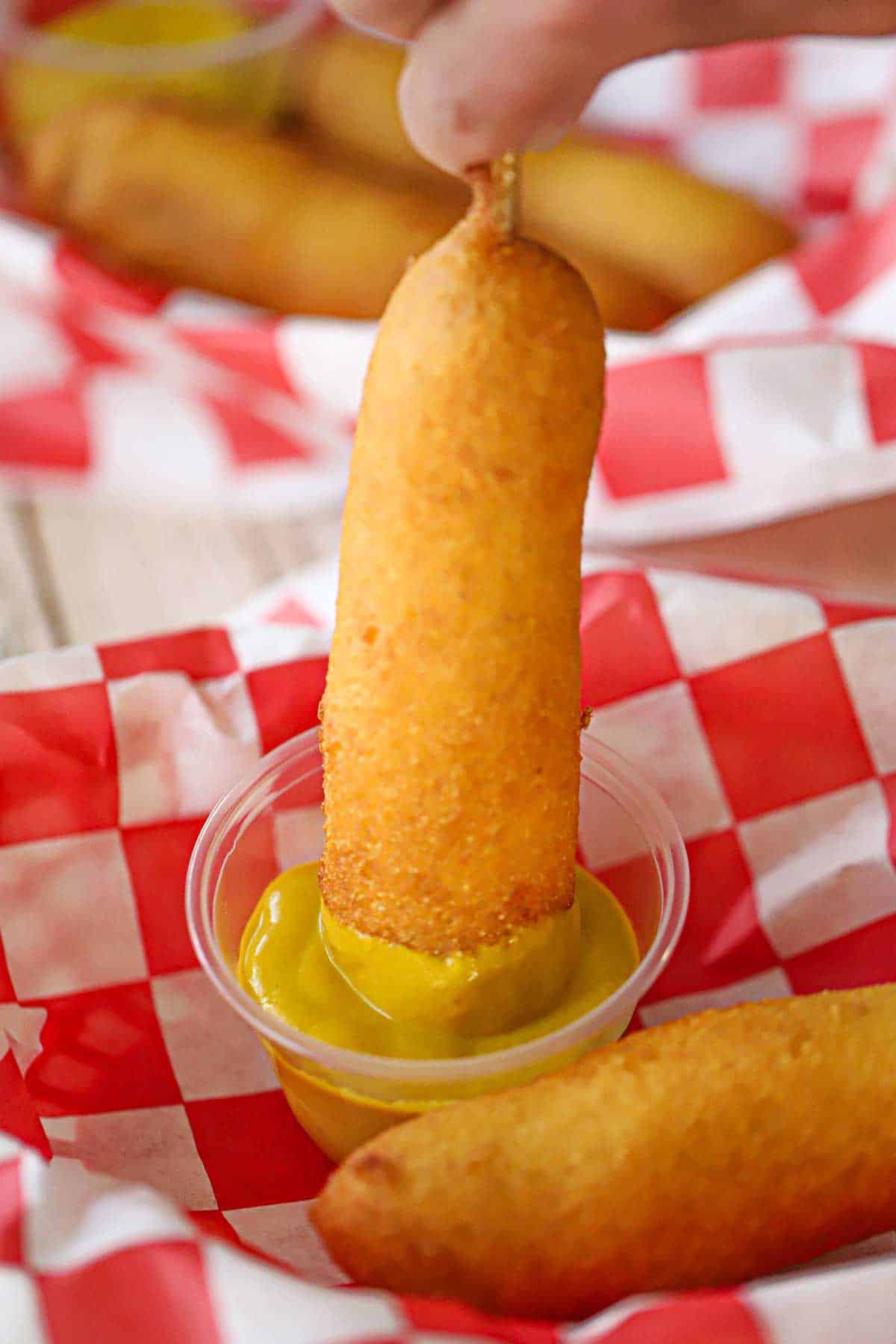 A person plunging a homemade corn dog into a small plastic container filled with yellow mustard.