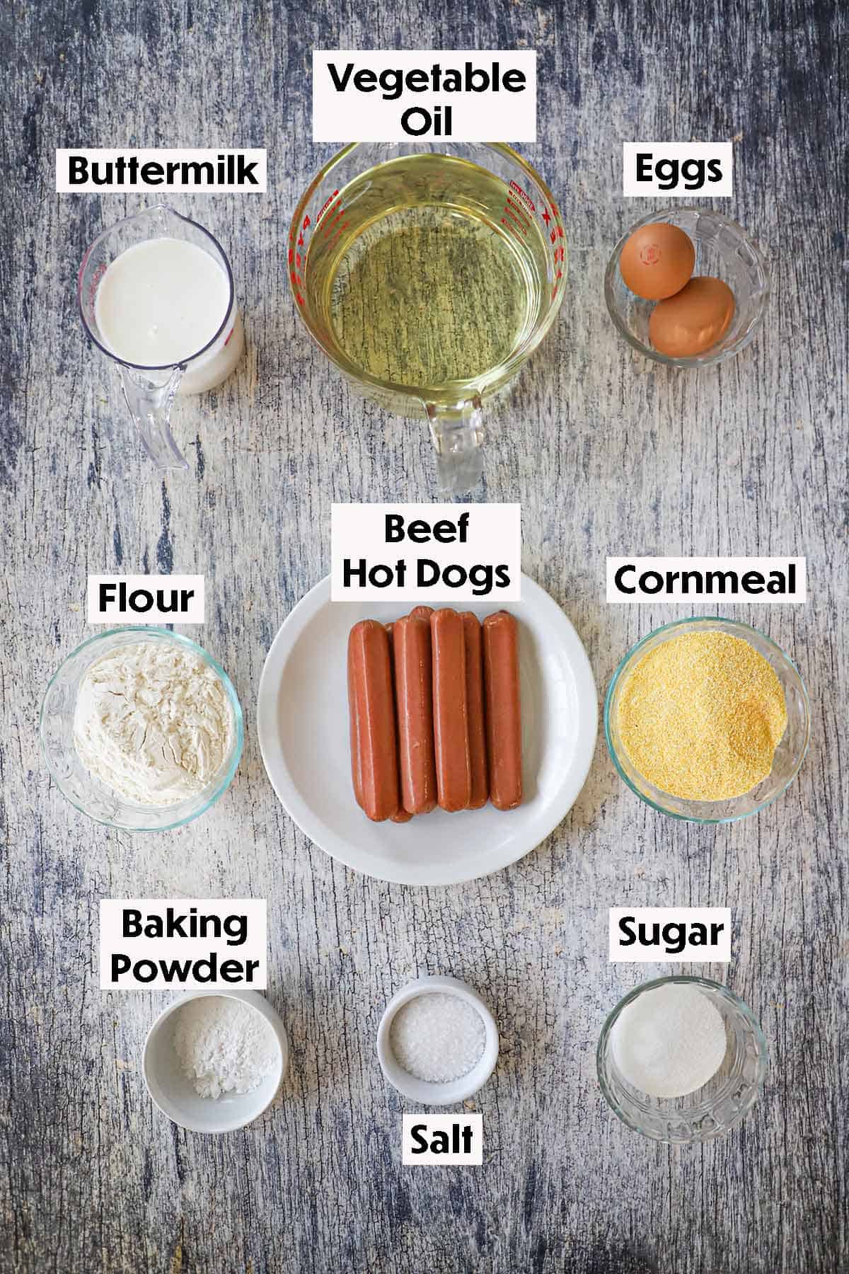 An arrangement of ingredients for homemade corn dogs on a grey wooden background including a pile of hot dogs, buttermilk, oil, eggs, flour, cornmeat, baking powder, salt, and sugar.
