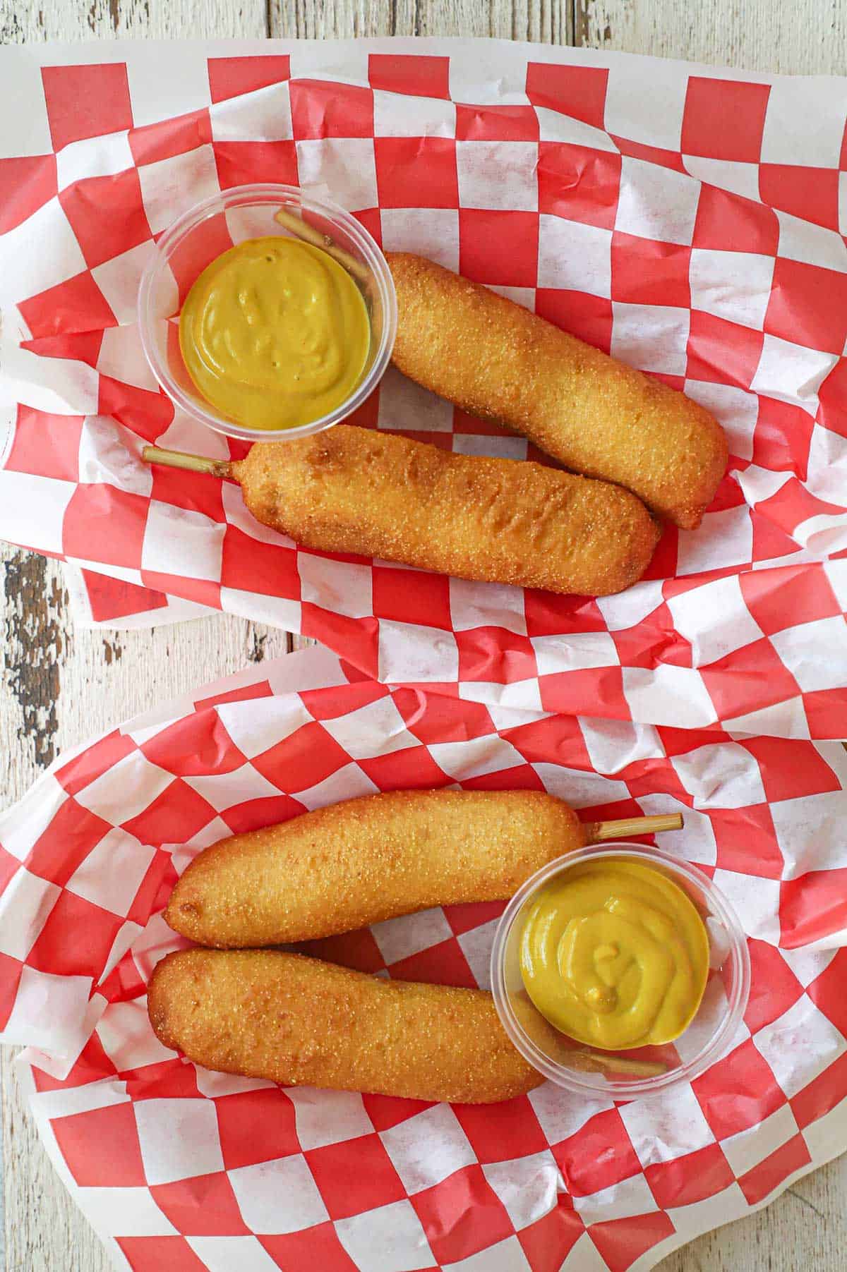 Two baskets lined with red and white checkered wax paper and filled each with two homemade corn dogs and a small container of yellow mustard.