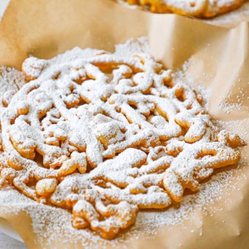 A close-up view of a homemade funnel cake topped with a dusting of powdered sugar and is resting on a brown piece of paper.