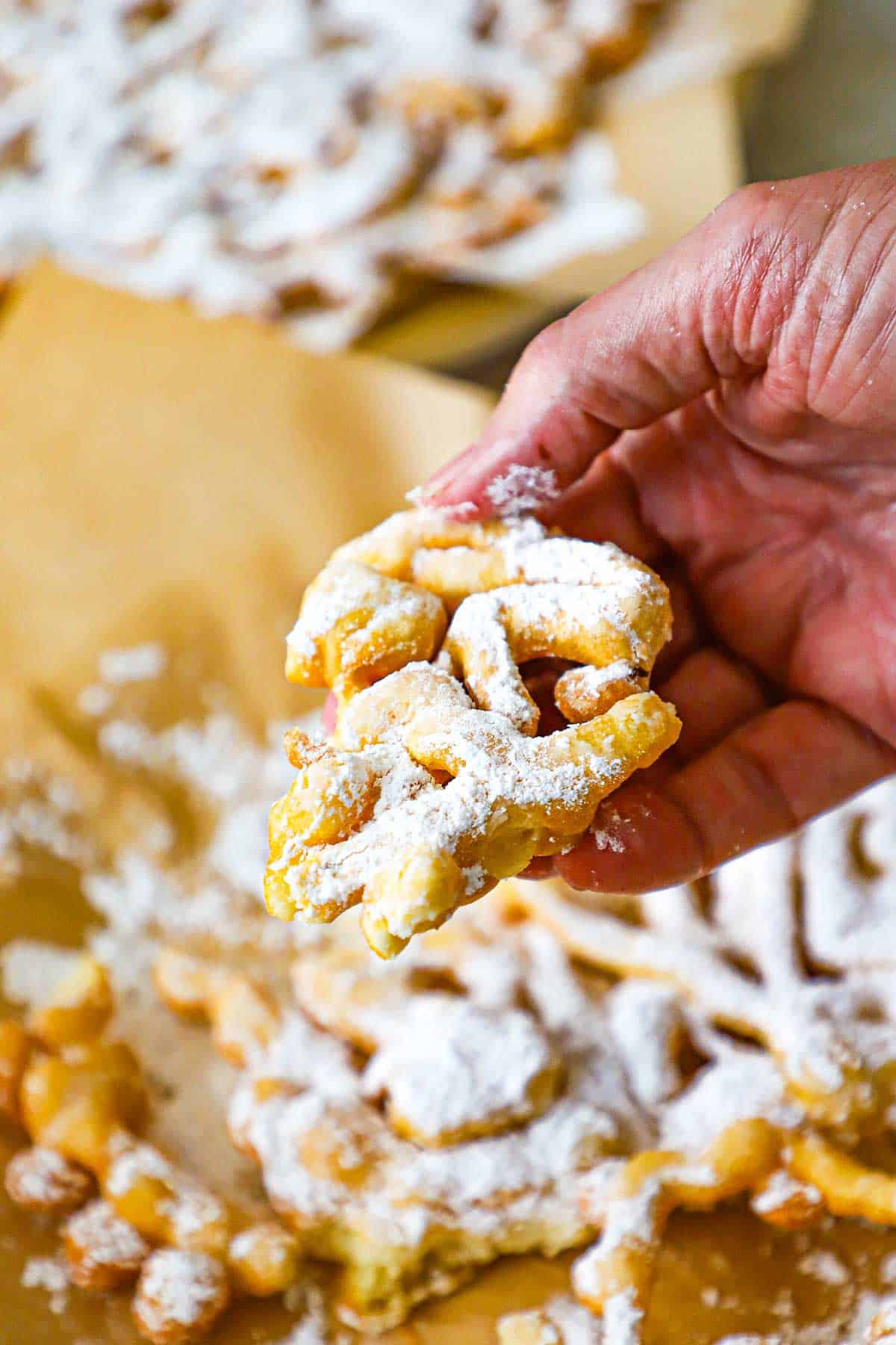 A person holding a portion of a homemade funnel cake in his hand over the rest of the funnel cakes below.
