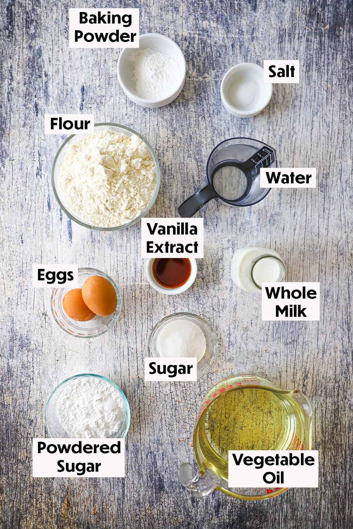 An arrangement of ingredients for homemade funnel cake on a grey wooden background including flour, baking powder, salt, water, eggs, vanilla, milk, sugar, oil, and powdered sugar.