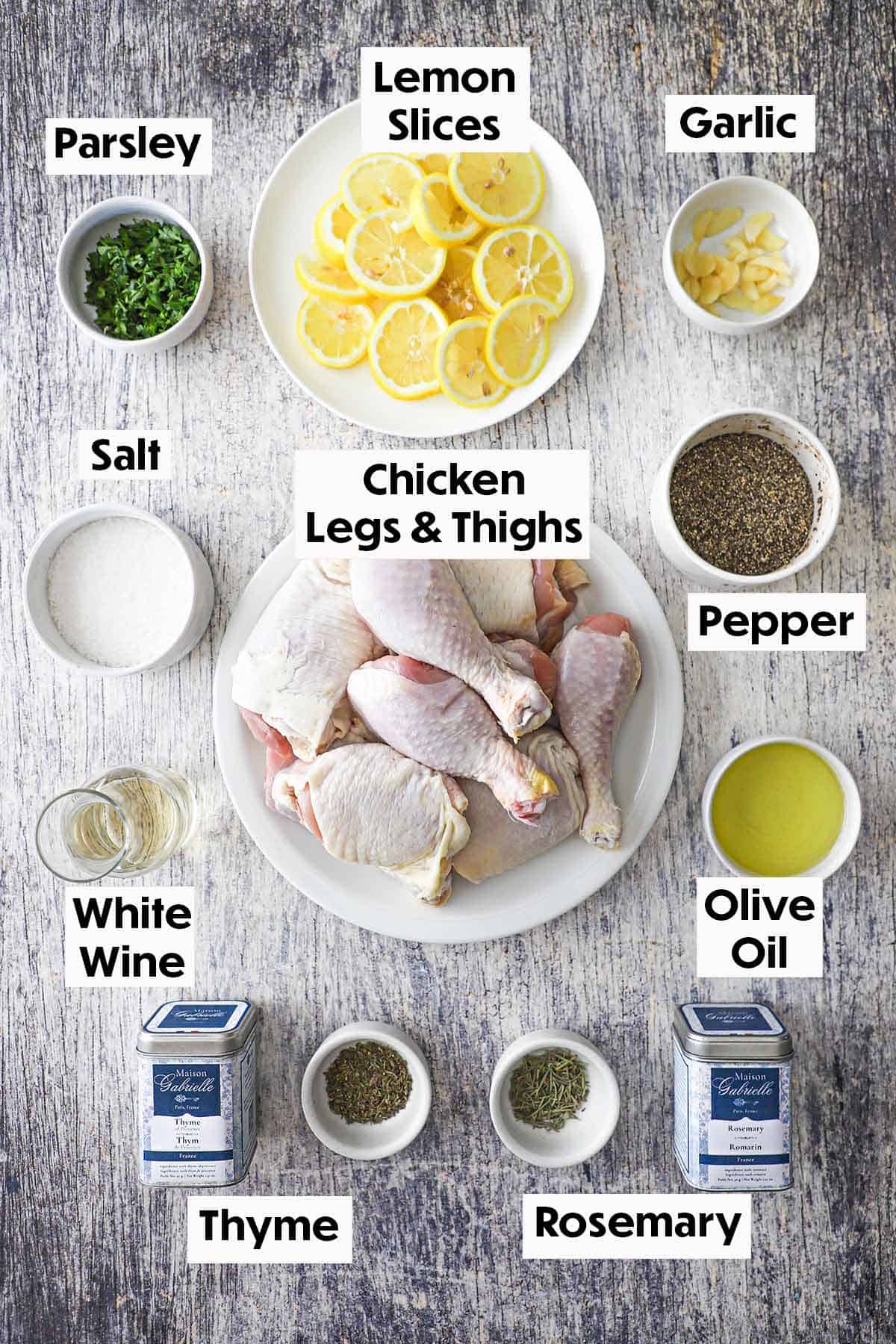 An arrangement of ingredients for Lemon Roast Chicken on a grey wooden background including uncooked chicken pieces, lemon slices, garlic, wine, oil, and seasonings. 