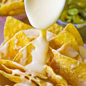 A spoon being used to drizzle easy homemade nacho cheese sauce onto rows of tortilla chips that are sitting side by side in a paper basket.