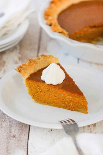 A slice of pumpkin pie on a white plate next to a whole pumpkin pie.