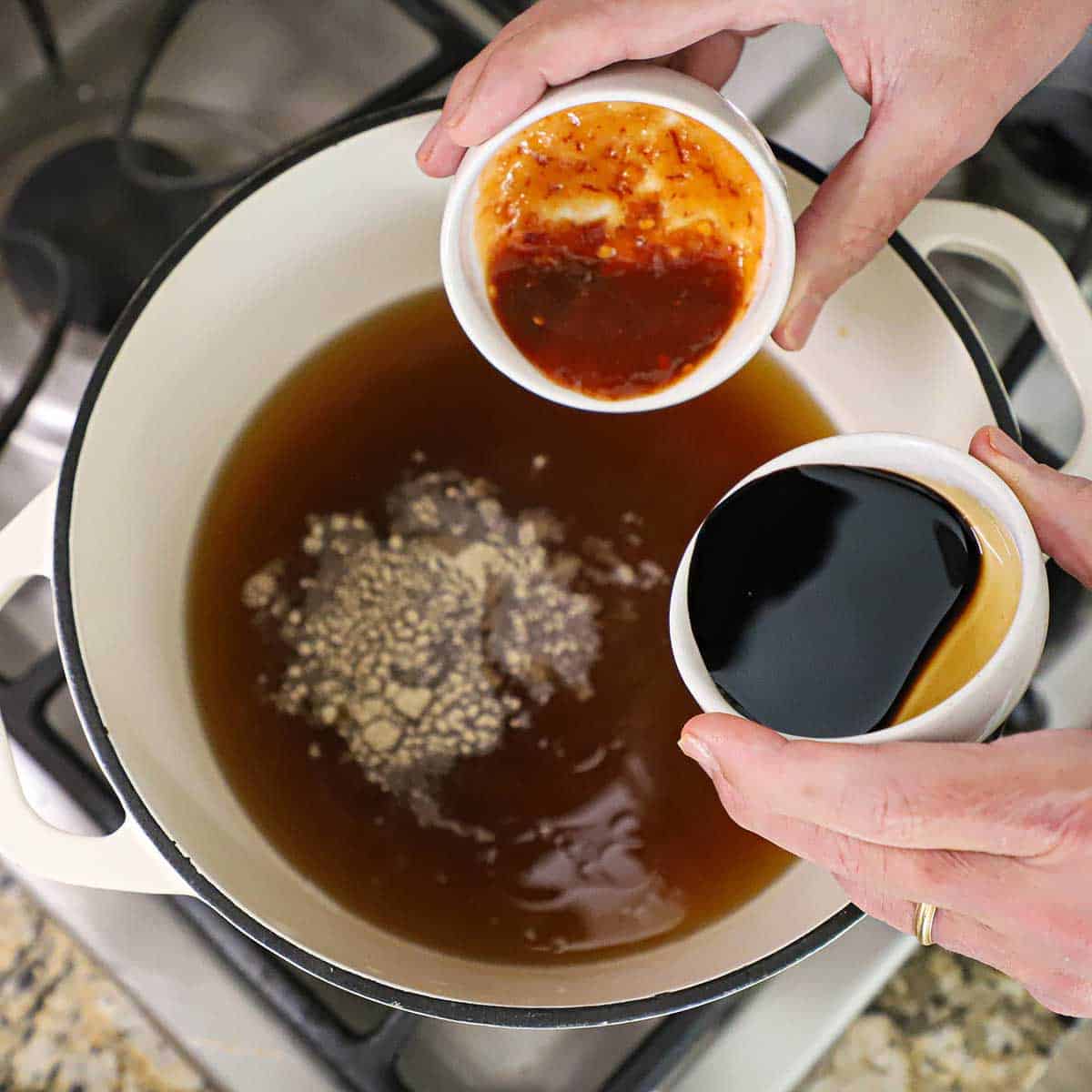 A person holding a small bowl filled with chili paste in one hand and the other hand holding a small bowl of black vinegar both over a pot filled with chicken broth and a sprinkling of white pepper.
