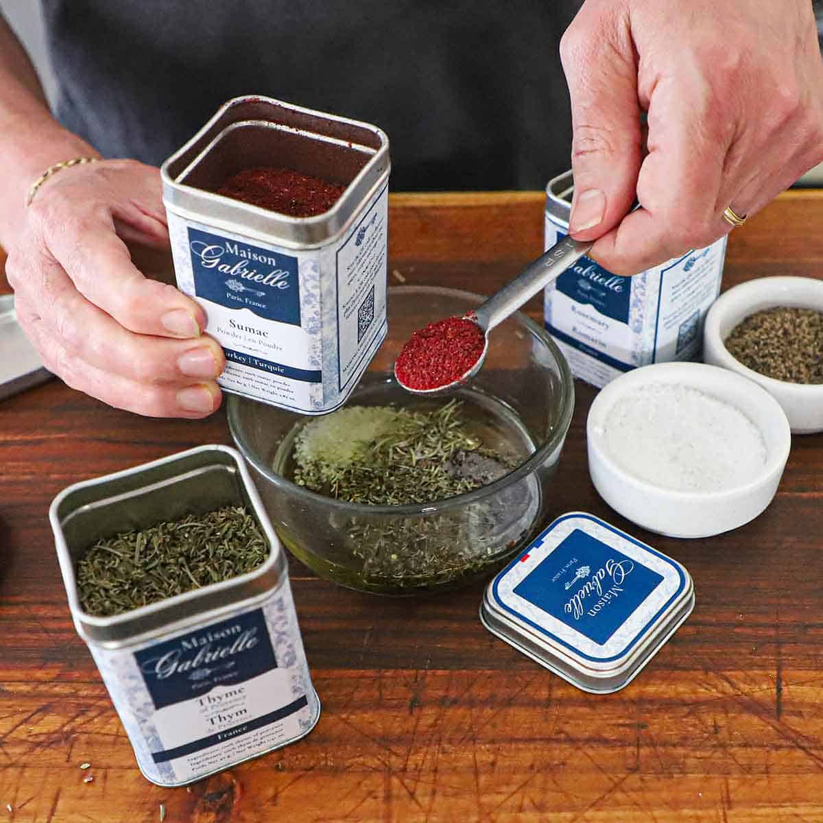 A person holding a teaspoon filled with sumac over a small glass bowl filled with olive oil, thyme, and rosemary.