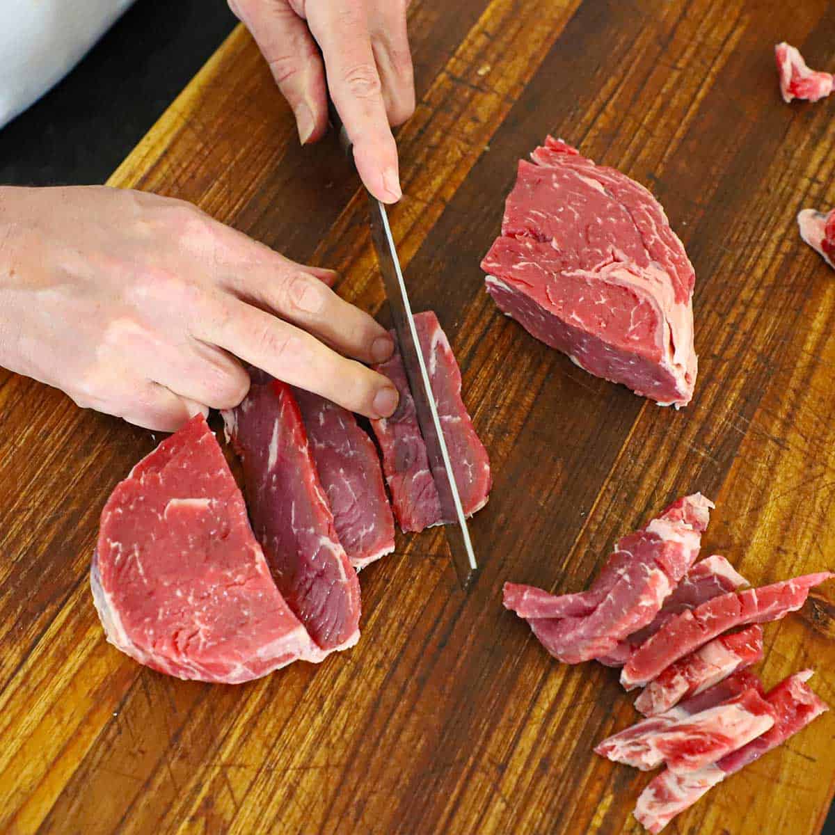 A person using a large Japanese knife to slice an uncooked and partially frozen ribeye steak into strips on a cutting board.
