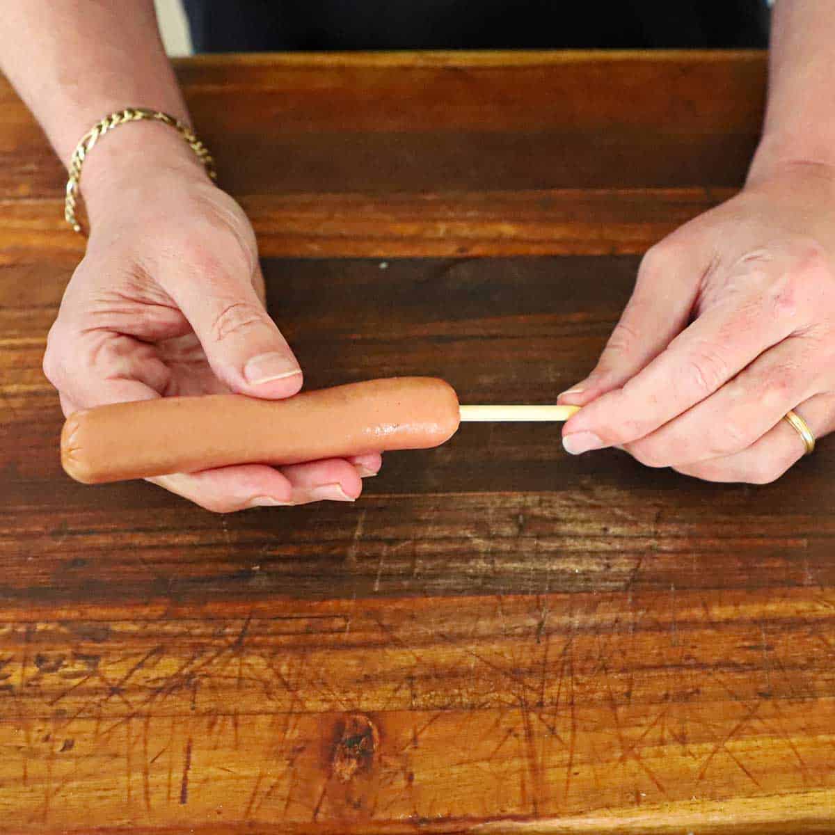 A person inserting a wooden skewer into the end of an uncooked beef hot dog over a wooden cutting board.