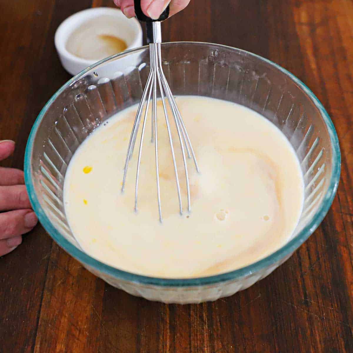 A person using a whisk to mix milk, water, eggs, and vanilla extract in a glass bowl on a wooden cutting board.