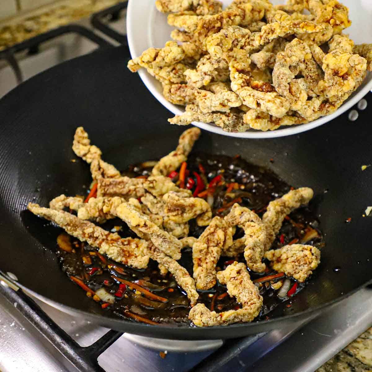 A person transferring a bowl-full of crispy strips of breaded steak into a wok that has a chili sauce and sautéed vegetables simmering in it.