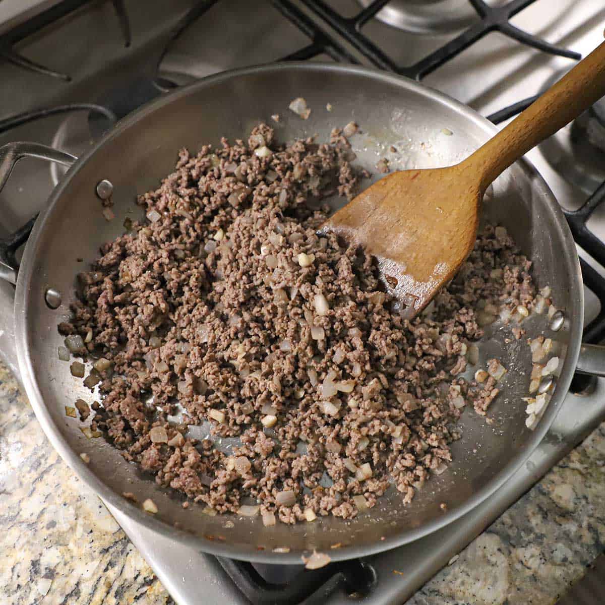 A large silver skillet filled with cooked ground beef that is mixed with chopped sautéed onions on gas stove.