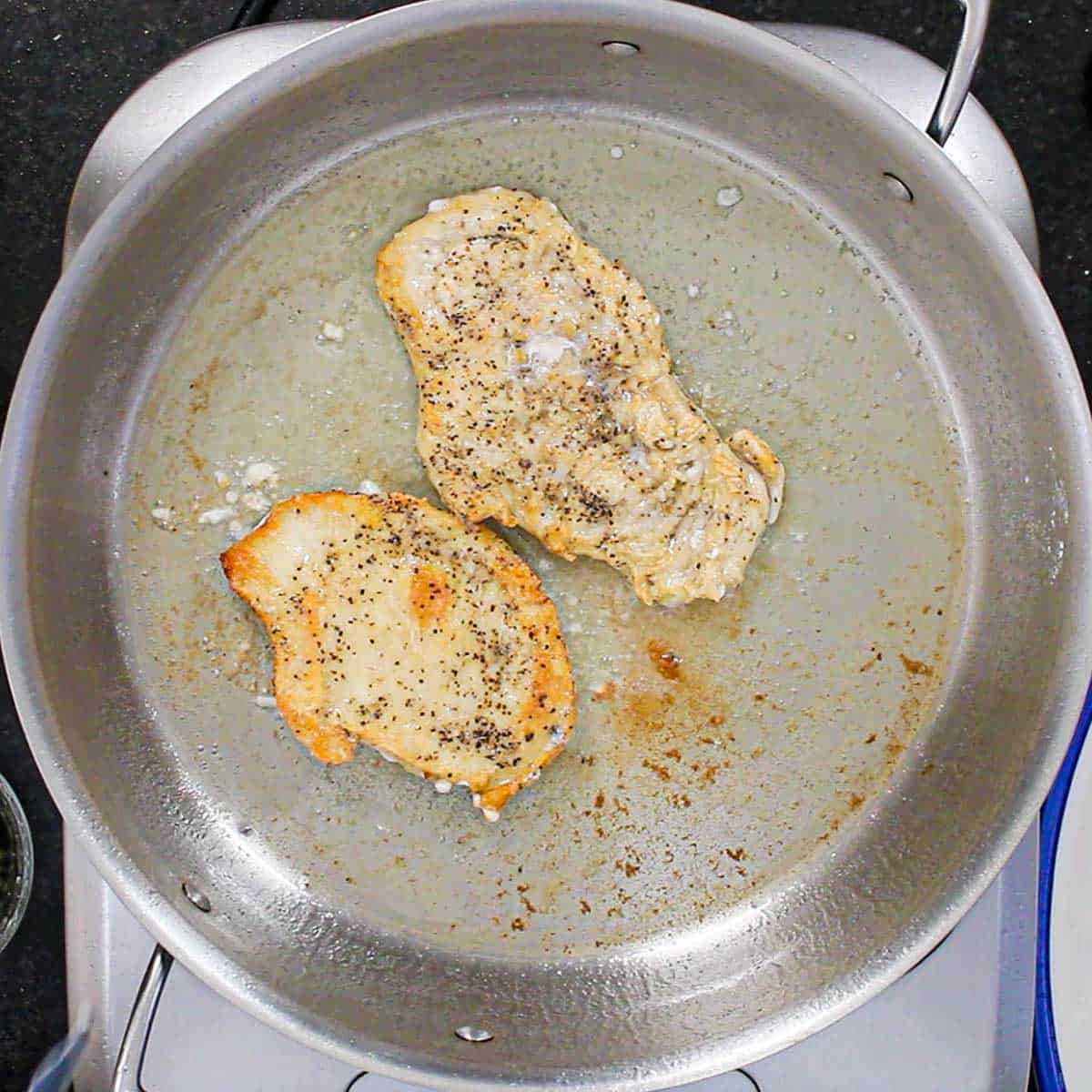 Two thin chicken breast cutlets being searing in olive oil in a large stainless steal skillet.