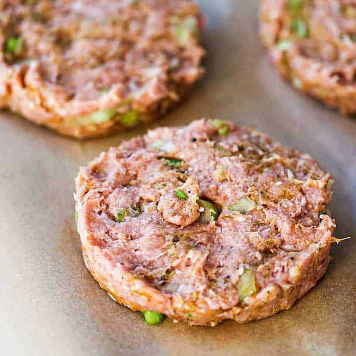 An uncooked curry turkey burger patty resting next to two more patties on a piece of brown paper.