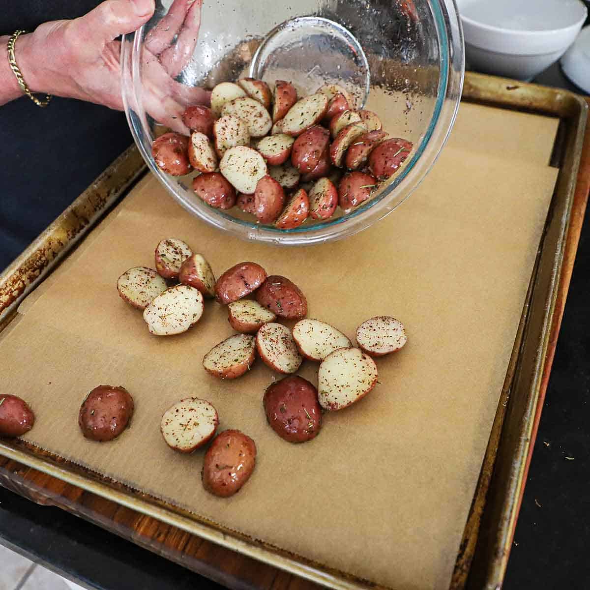A person transferring oiled and seasoned halved baby red potatoes on a large baking sheet that is lined with parchment paper.