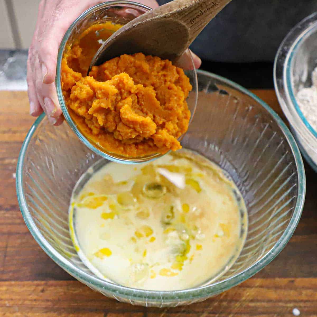 A person using a wooden spoon to transfer pumpkin purée from a small glass bowl into a large bowl filled beaten eggs, milk, and oil.
