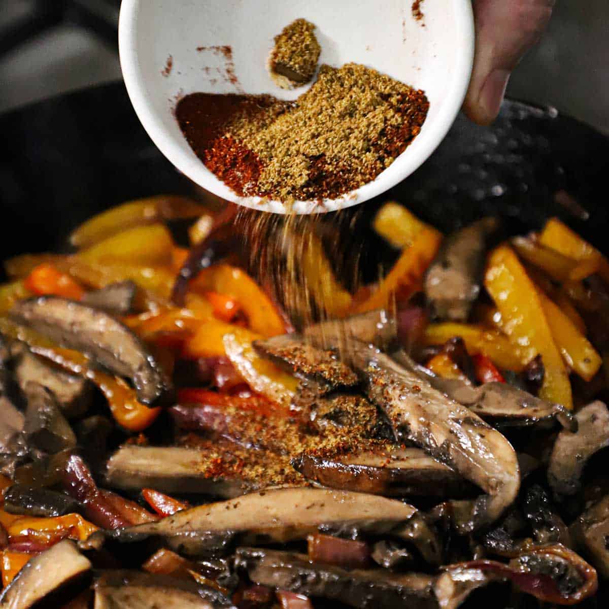 A person transferring dried spices from a small white bowl into a skillet that is filled with sautéed strips of portobello mushrooms, bell peppers, and onions.