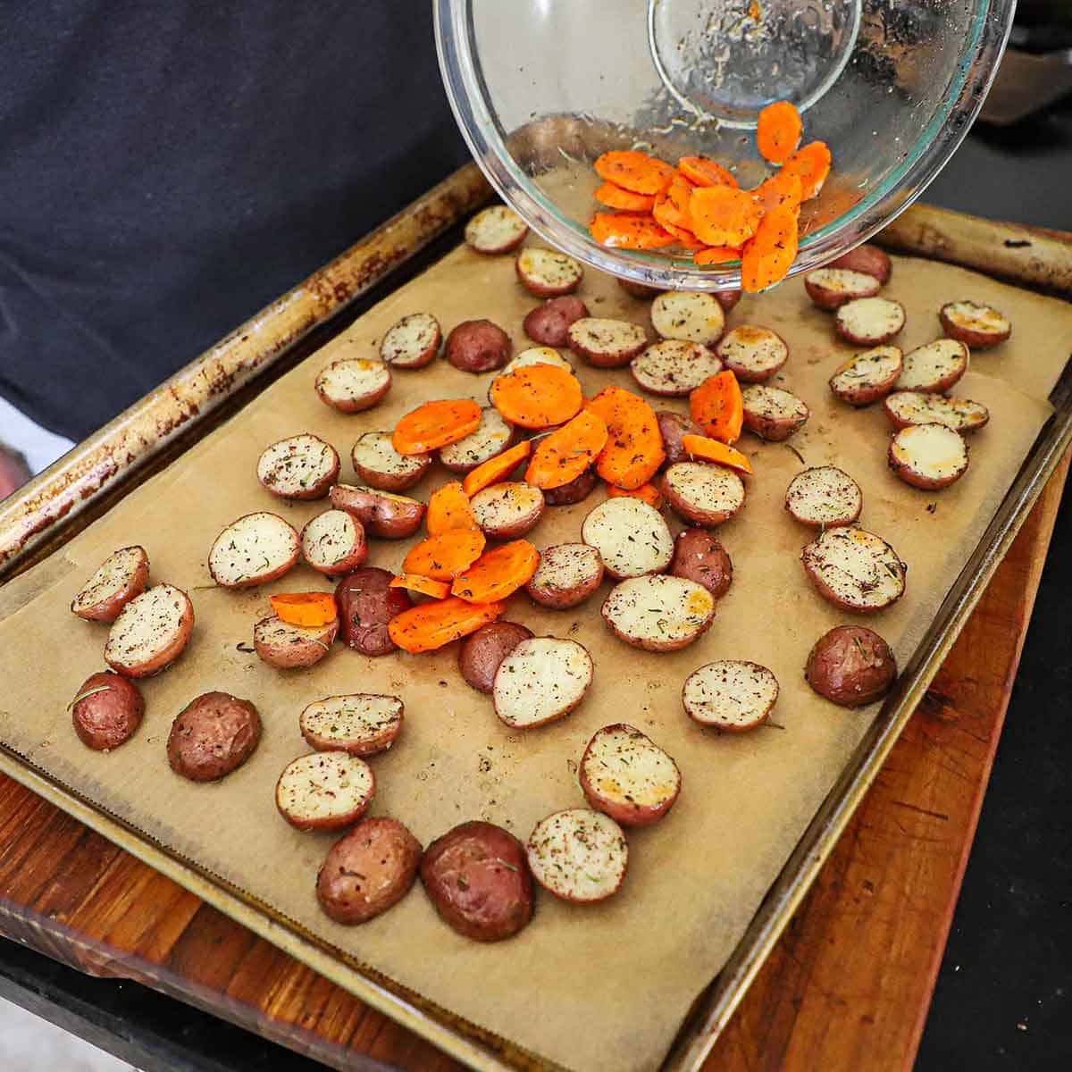 A person transferring slices of oiled and seasoned carrots onto a baking sheet that is lined with parchment paper and has lightly toasted baby red potatoes on it.