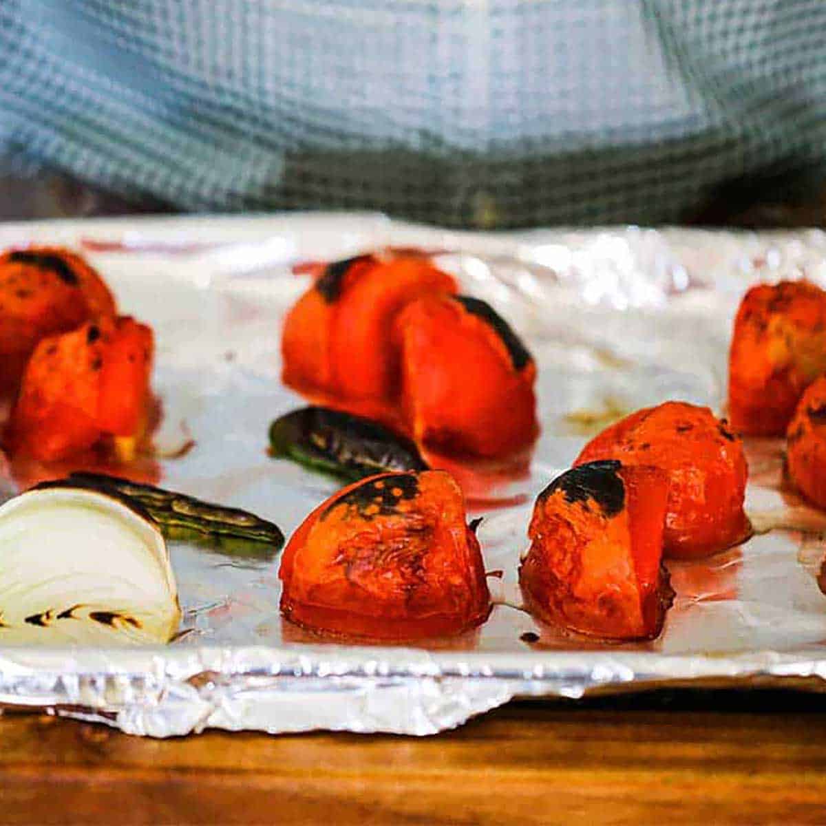 A person holding a baking sheet lined with aluminum foil and is holding roasted tomatoes, peppers, and white onion.