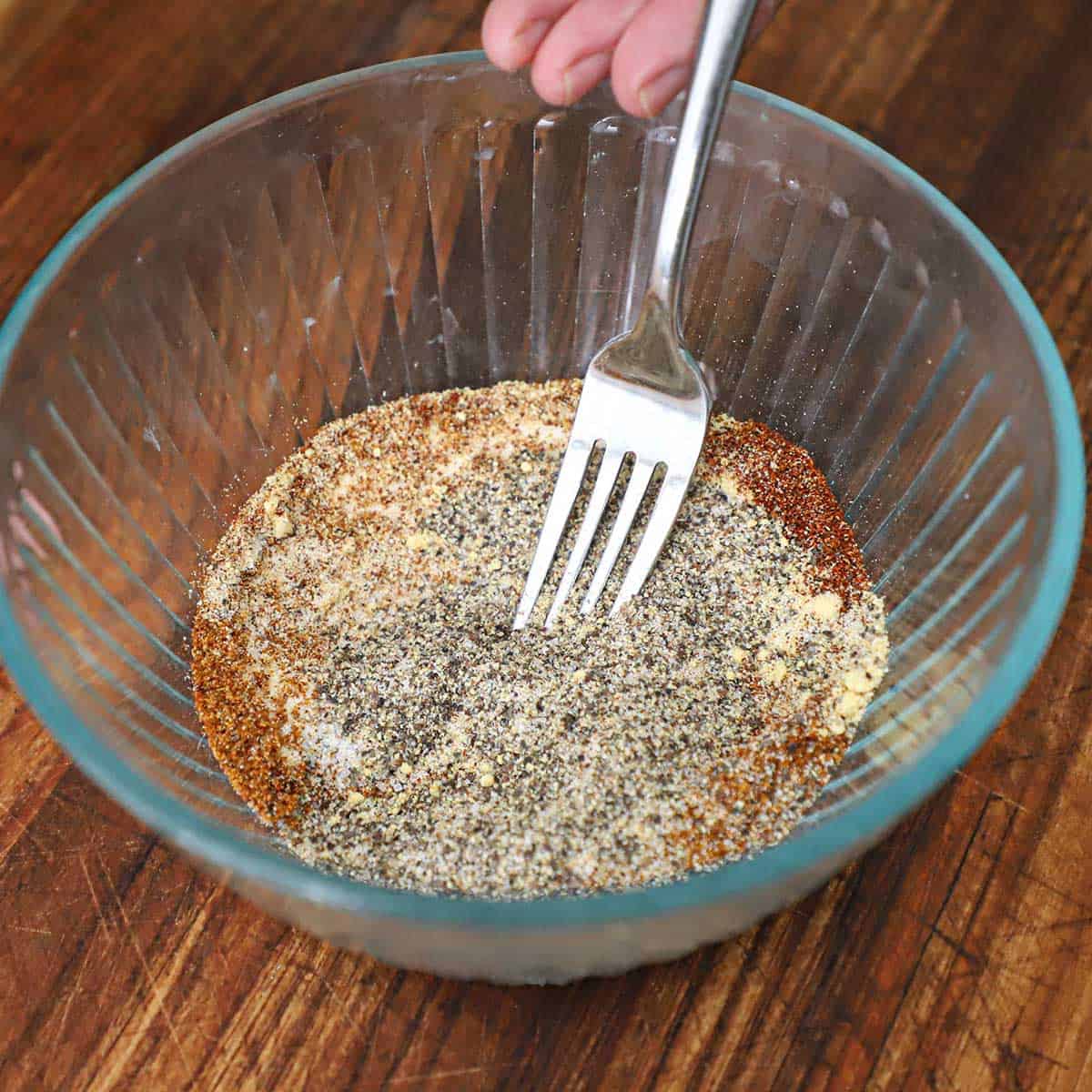 A person mixing a best beef rub in a small glass bowl on a wooden cutting board.