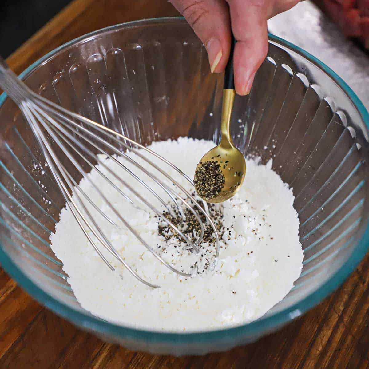 A person transferring ground black pepper from a small gold spoon into a glass bowl filled with cornstarch with a whisk resting in the middle of the bowl.