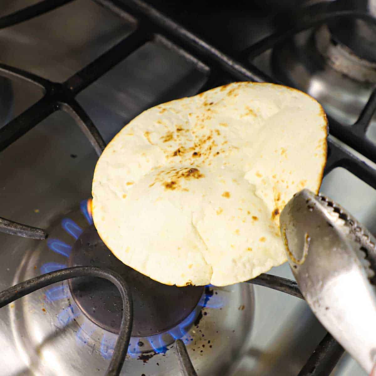 A person using a pair of metal tongs to lightly char a flour tortilla over a gas flame on a gas stove.