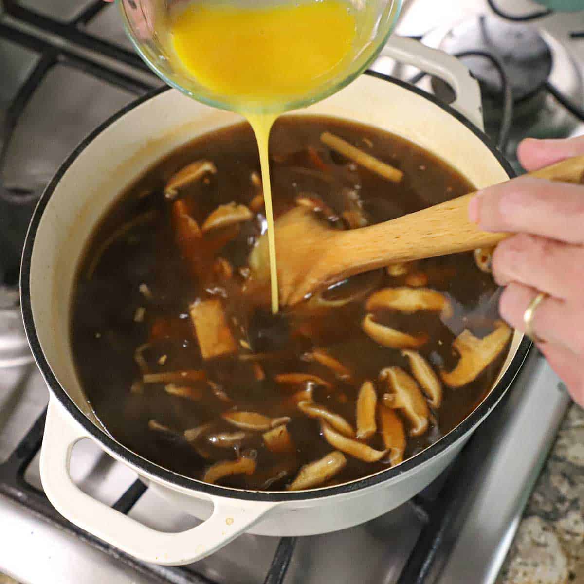 A person stirring in two lightly beaten eggs from a glass bowl into a large pot filled with simmering homemade Chinese hot and sour soup.
