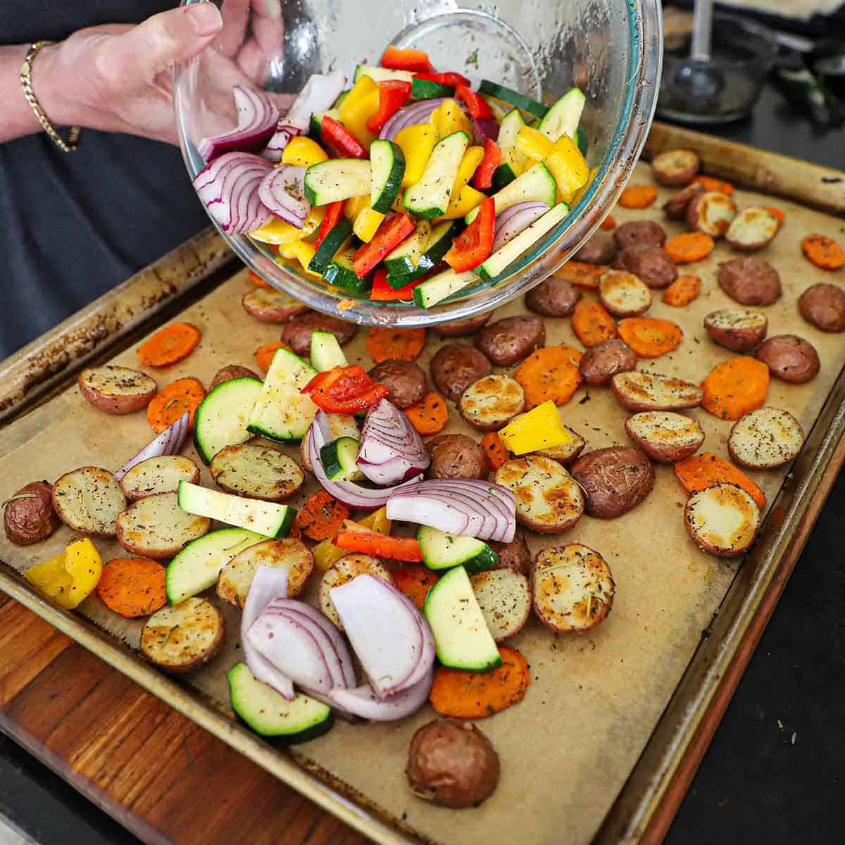A person transferring sliced zucchin, red onion wedges, and strips of bell peppers onto a baking sheet that is filled with toasted baby red potatoes and carrot slices.