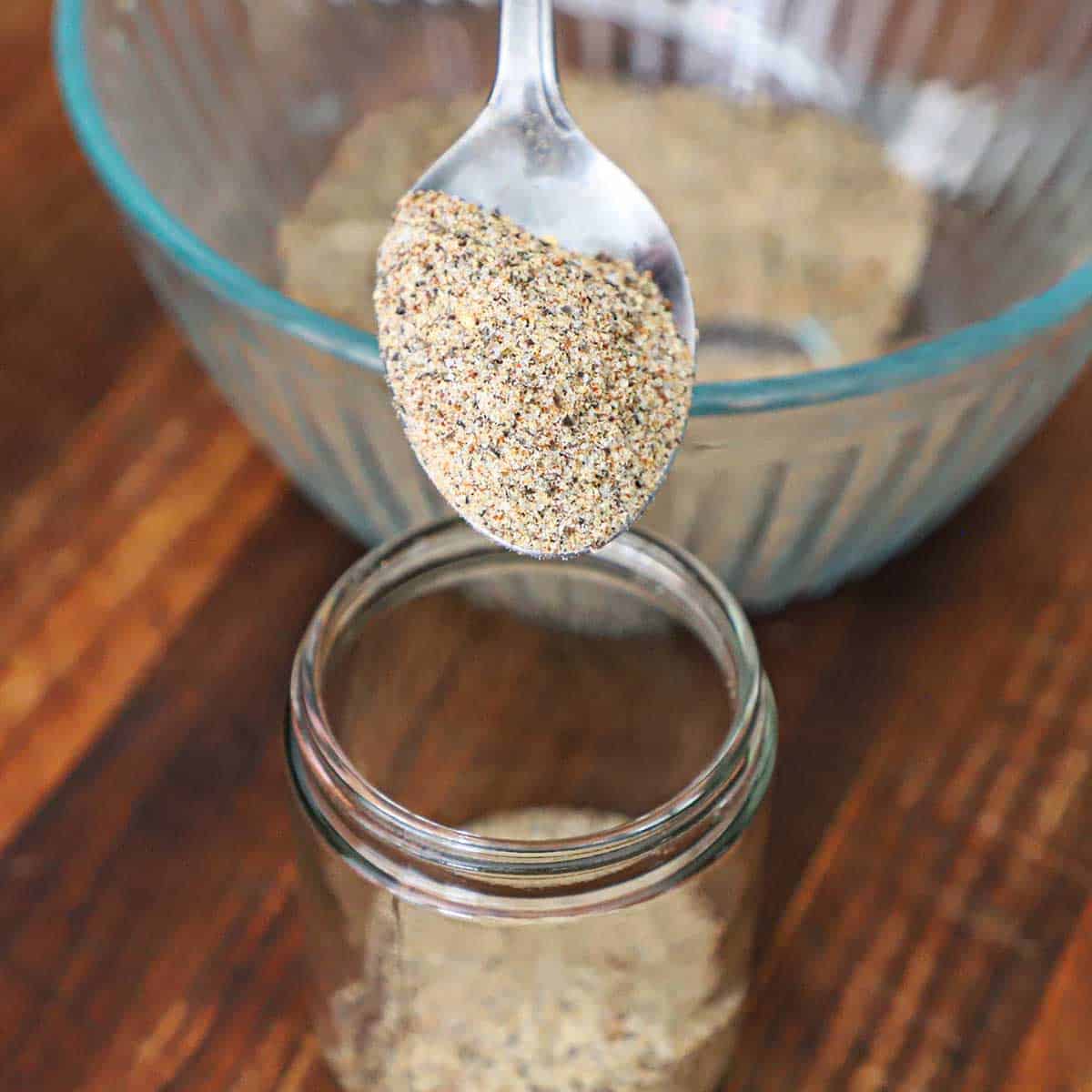 A person using a silver spoon to transfer Best Beef Rub from a glass bowl into a small glass jar.