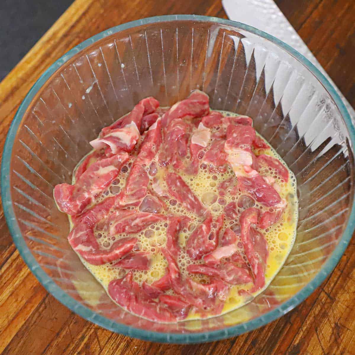 Strips of uncooked ribeye steak resting in a bowl that has two beaten eggs in it.