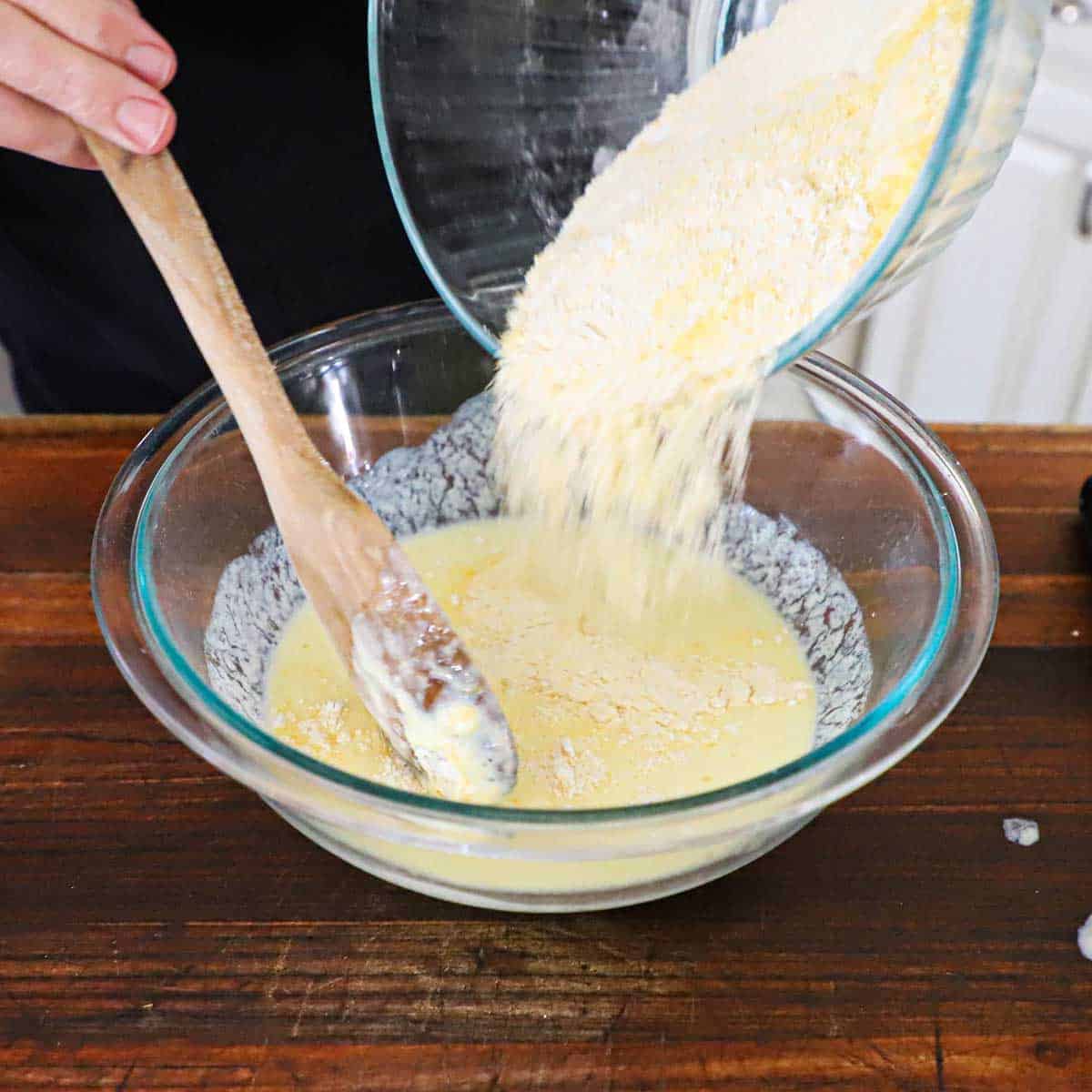 A person transferring a dry flour and cornmeal mixture from one bowl into another glass bowl filled with an egg and buttermilk wet mixture.
