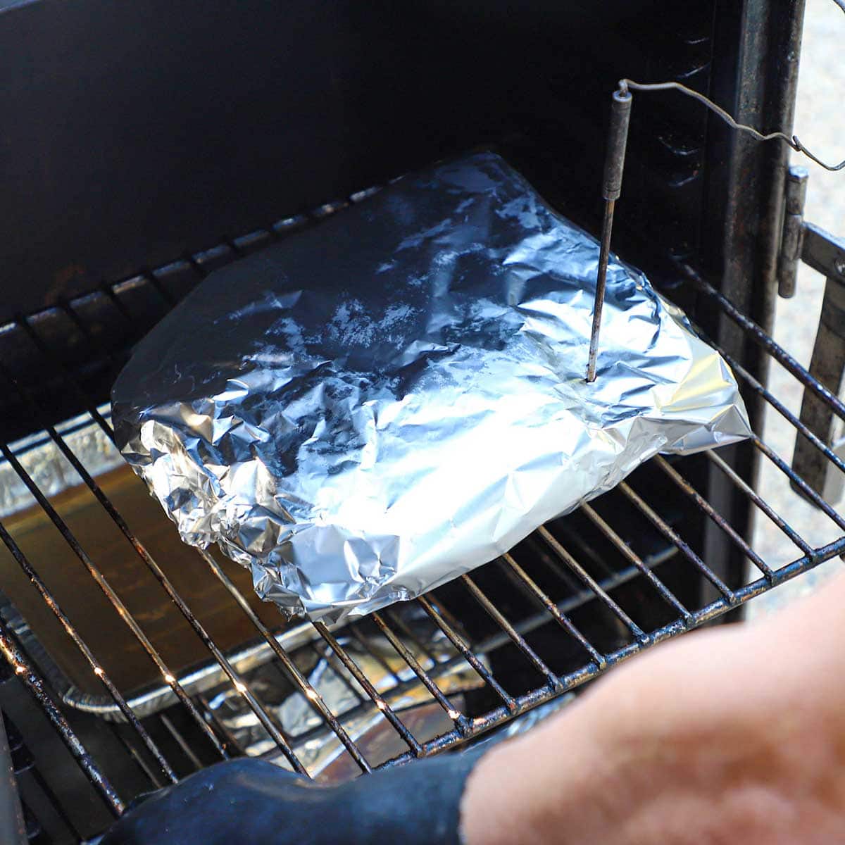 A person pulling a grate from an electric smoker that is holding a beef brisket flat that has been tightly wrapped in foil with a digital thermometer piercing through the foil into the meat.