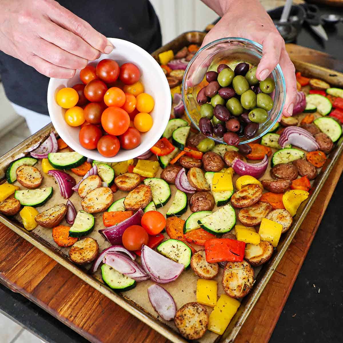 A person transferring a bowl filled with cherry tomatoes in one hand and a bowl of olives in the other hand onto a baking sheet filled with Mediterranean roasted vegetables on it.