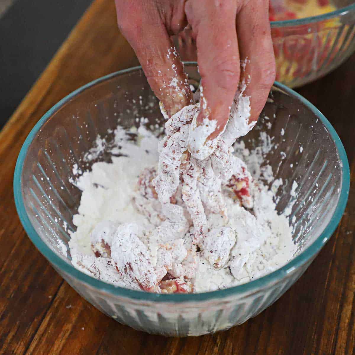 A person using his hands to coat egg-soaked beef strips with seasoned cornstarch in a glass bowl.