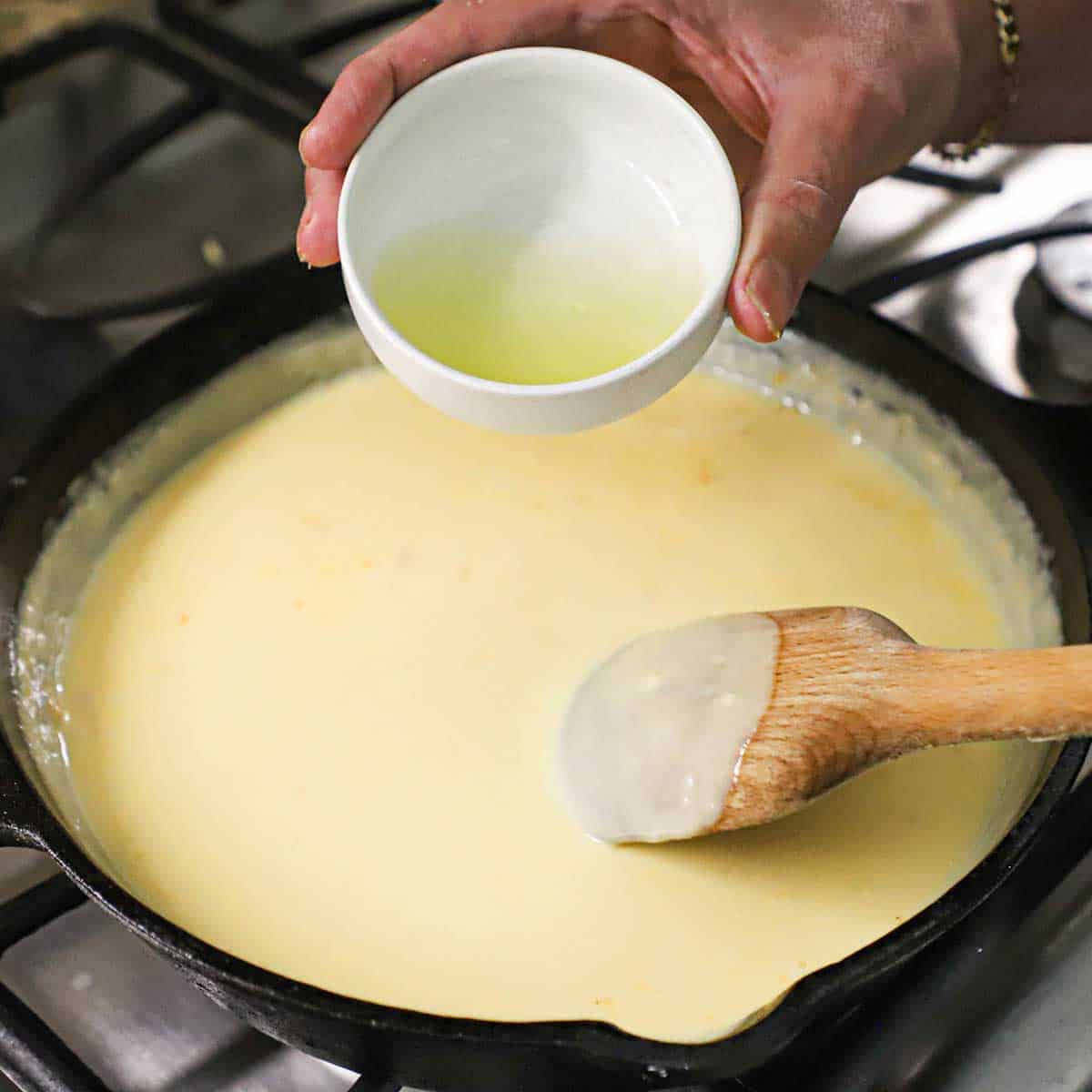 A person getting ready to transfer a small bowl of pickled Jalapeño brine into a skillet that is filled with a simmering nacho cheese sauce.