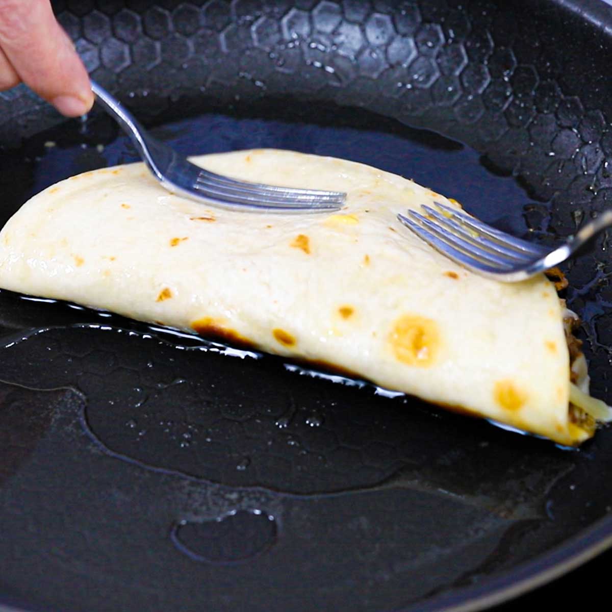 A person using two forks to hold down a taco quesadilla that is being lightly fried in a non-stick skillet with a layer of shimmering oil in it.