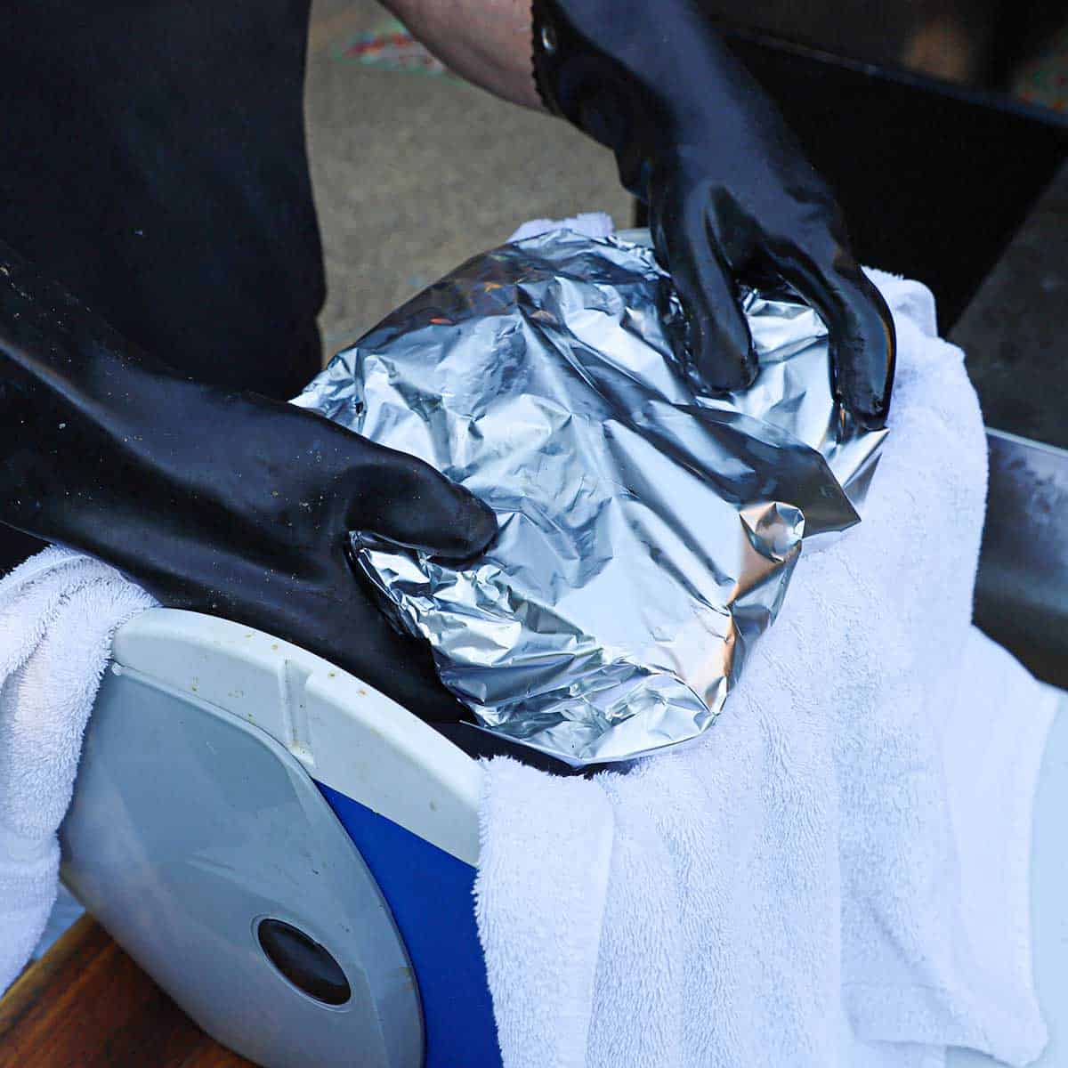 A person placing a fully cooked brisket that is still wrapped in foil into an Igloo cooler that is lined with a white towel.