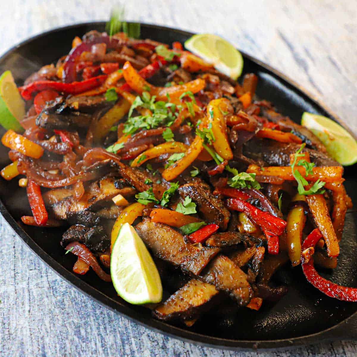 A close-up view of veggie fajitas that are sizzling on a hot fajita skillet with lime wedges sitting on the side of the pan.