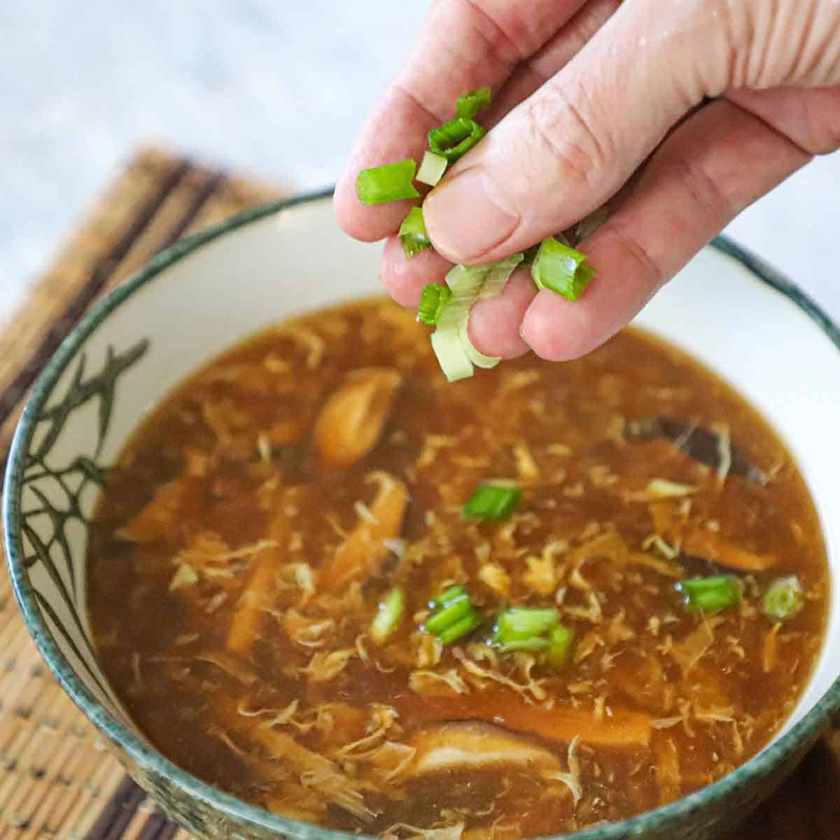 A person sprinkling chopped scallions over the top of a bowl filled with steaming homemade Chinese hot and sour soup.