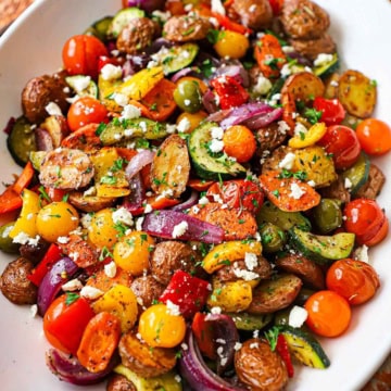 A close-up view of a large white platter filled with Mediterranean roasted vegetables garnished with crumbled feta cheese and chopped Italian parsley.