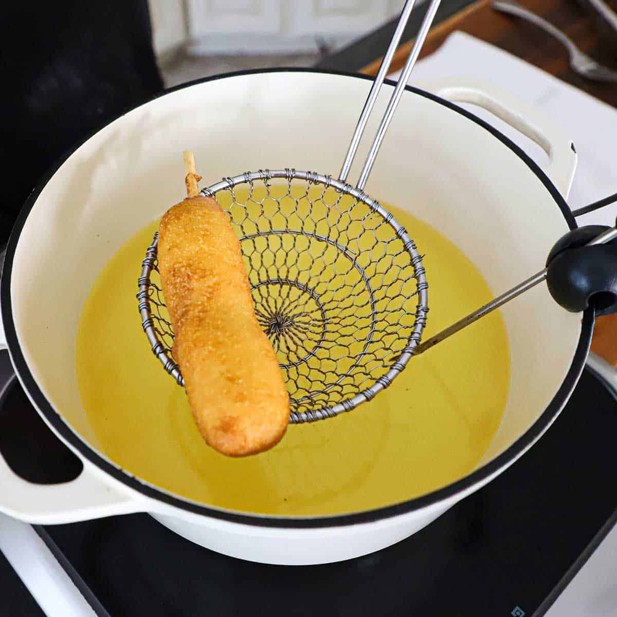 A person using a metal spider to lift a freshly deep-fried corn dog from a white pot filled with hot oil.