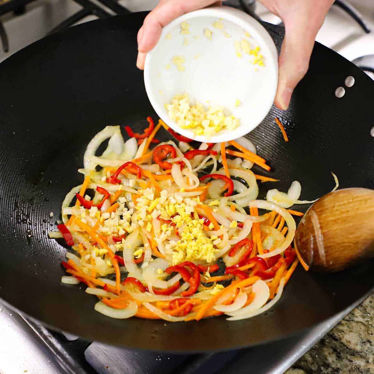 A person transferring mince garlic and minced ginger from a small bowl into a wok that has stir-fried onions, carrots, and peppers in it.
