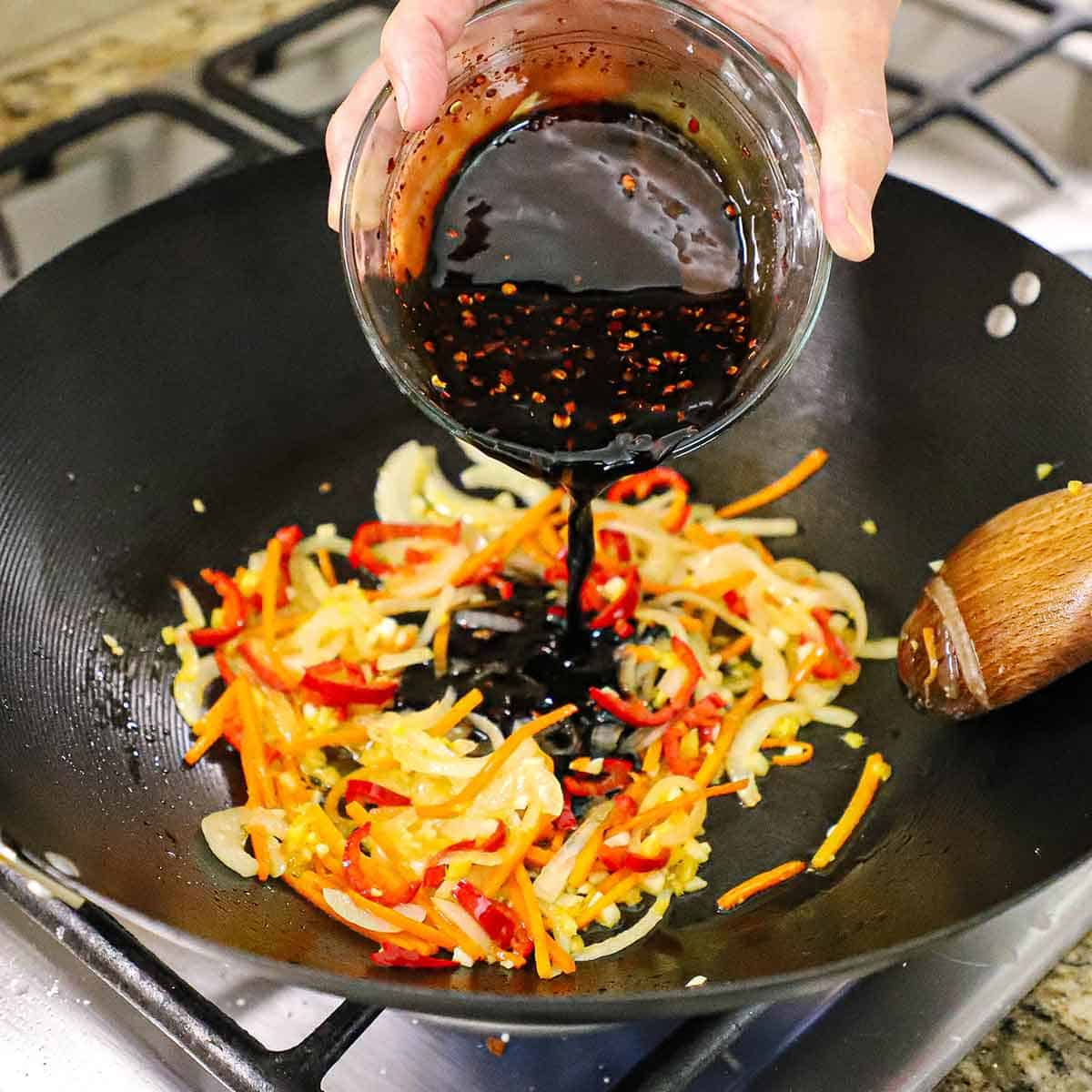 A person pouring an Asian sweet and spicy sauce from a glass bowl into a wok that has simmering vegetables that are being stir-fried with the sauce.